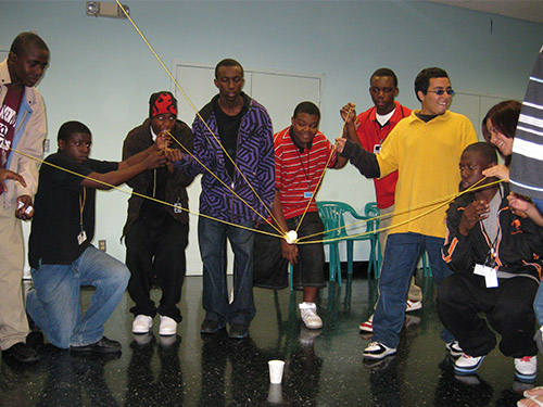 A group of participants work together to manage a string system with the goal of holding up a ball in the center of the room.